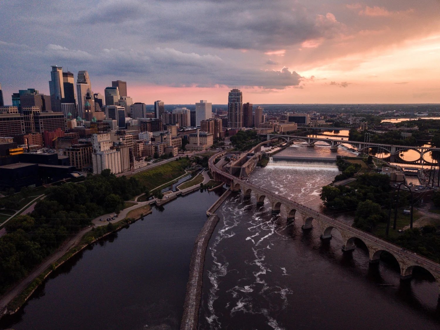 Aerial view of Minneapolis skyline at sunset with rivers, bridges, and downtown buildings under a colorful pink and purple sky